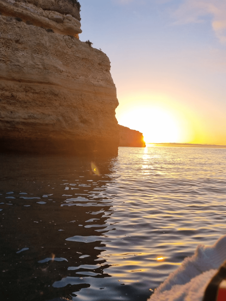 Sunset over calm sea with rocky cliffs on the left.