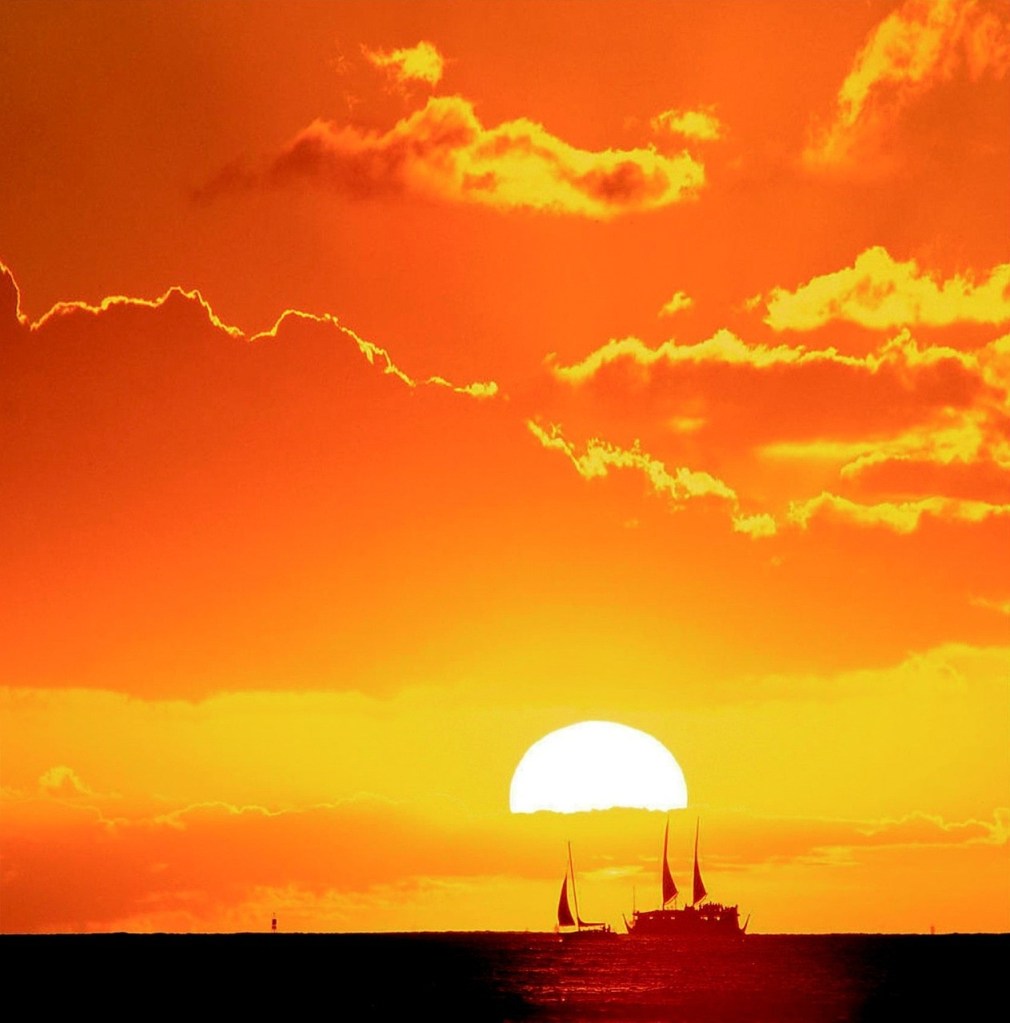 Silhouetted sailboats on ocean against vibrant orange sunset.