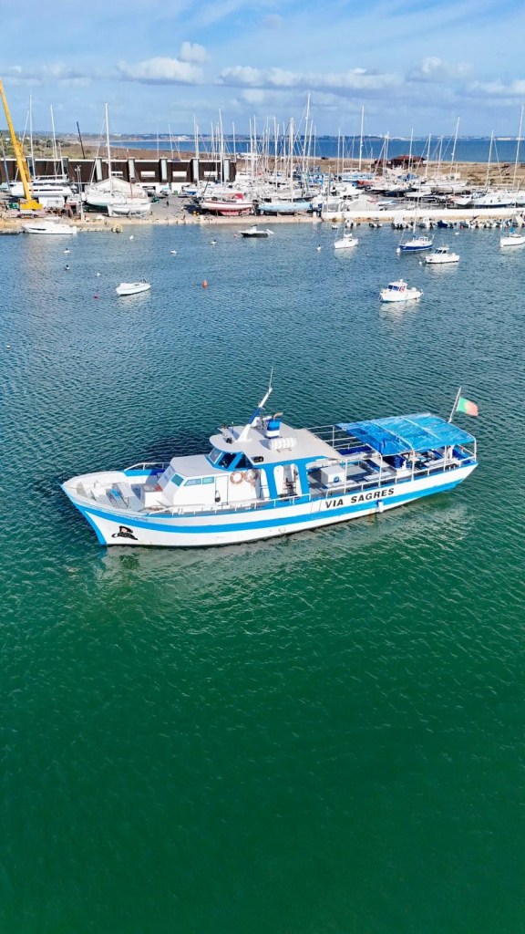 Boat named Via Saggese in a marina with sailboats docked in the background.