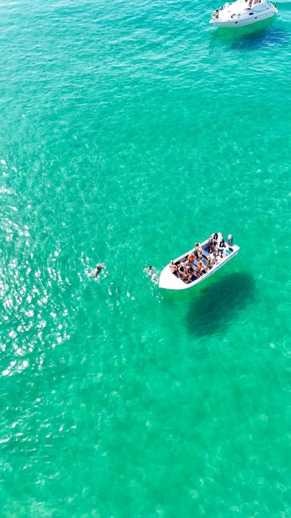 Boats on clear turquoise water with people swimming nearby.
