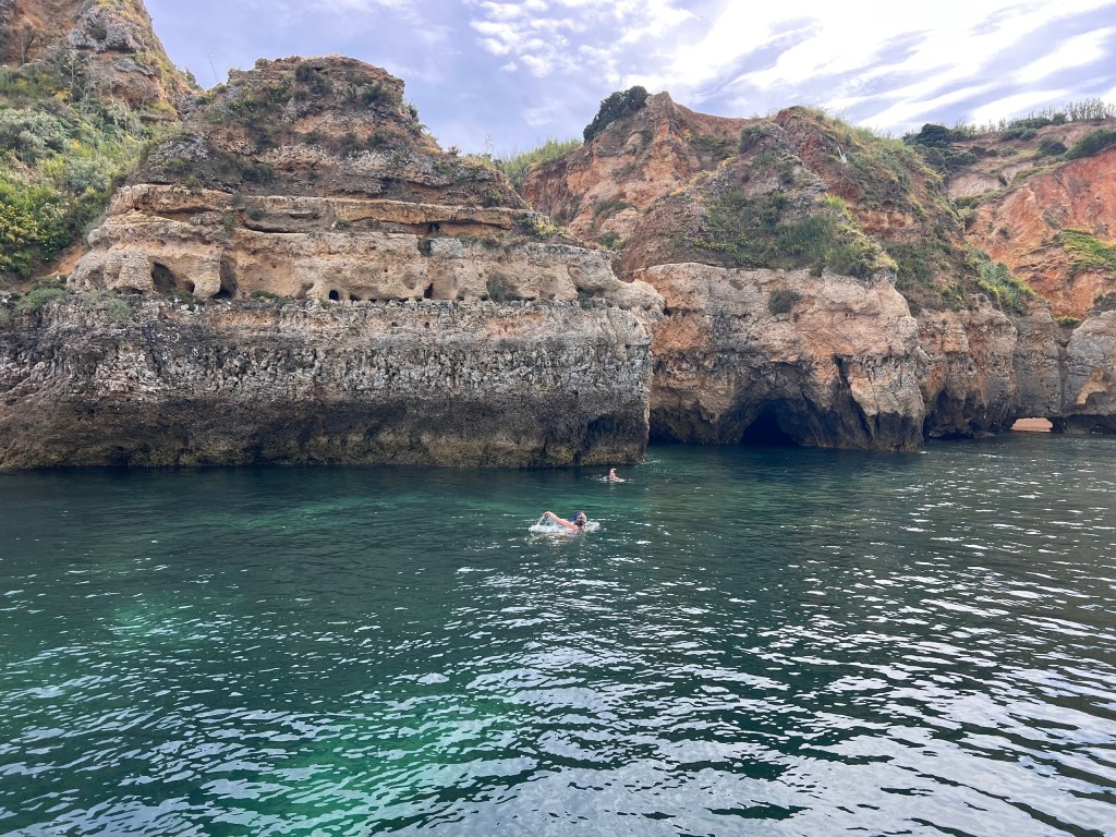 Swimmers near rocky cliffs and caves in a calm sea.