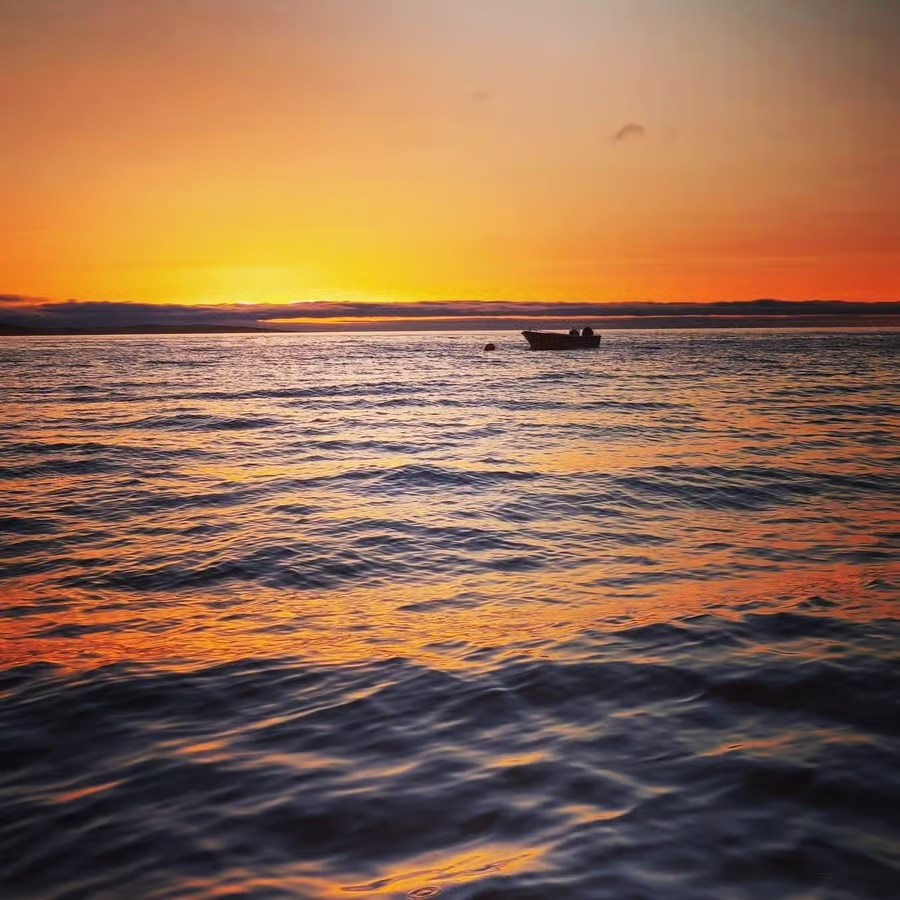Boat on ocean at sunset with orange sky and calm waves.