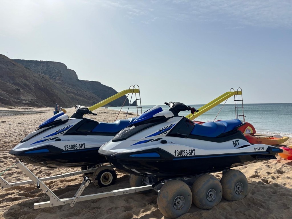 Two jet skis on trailers at a sandy beach with a rocky hill.