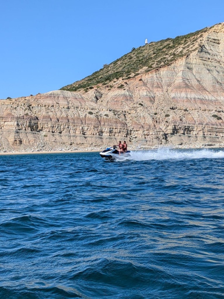 Two people riding a jet ski on blue water near a rocky cliff.