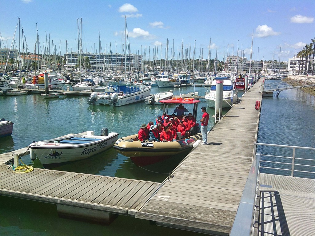 People in red jackets on a boat at a marina with numerous docked sailboats.