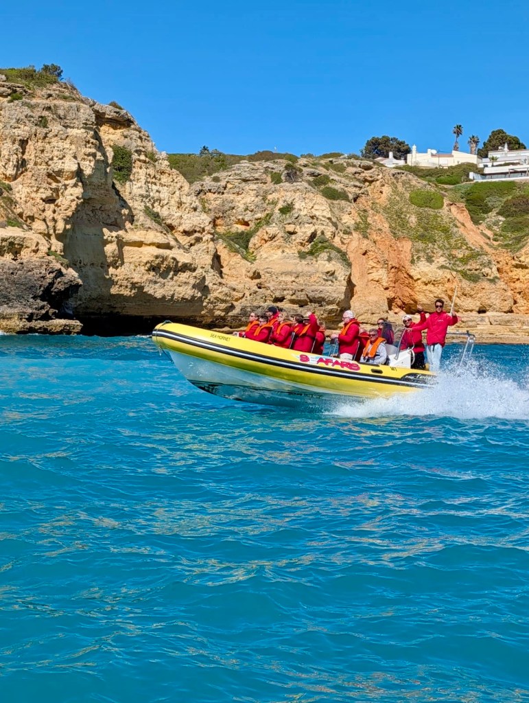 Yellow inflatable boat with people in life vests on blue water near rocky cliffs.