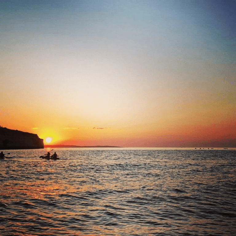 Sunset over ocean with silhouettes of kayakers and distant cliff.