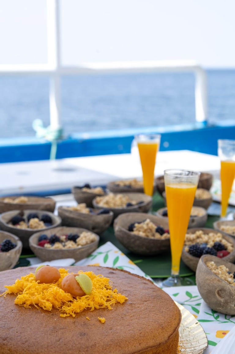 Cake with toppings and fruit bowls with orange drinks on a table by the sea.
