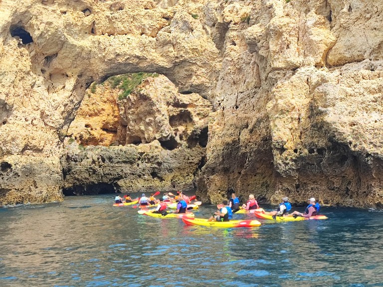Group kayaking near rocky cliffs and natural arch on calm water.