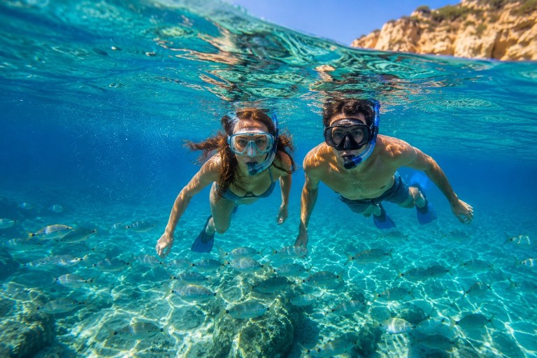 Two people snorkeling underwater near rocky shore with clear blue water.