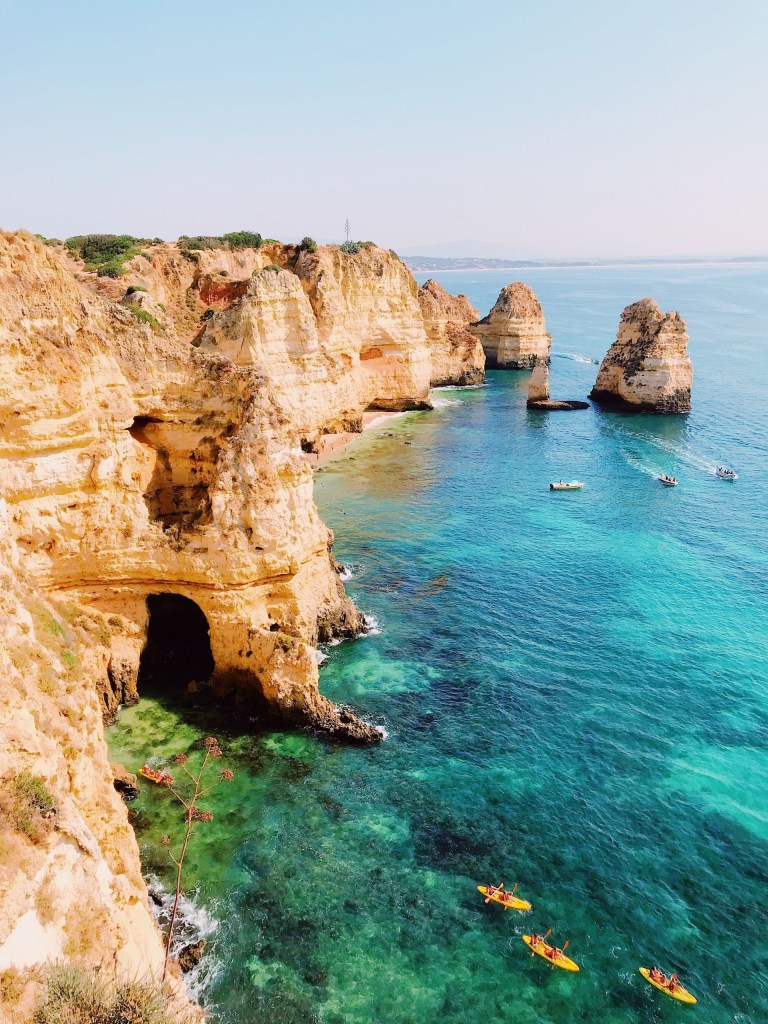 Cliffside with clear turquoise water, boats, and rock formations in a sunny coastal scene.